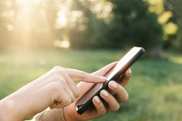 Photo of Closeup of female hands using a smart phone, nature background. Woman using mobile phone in sunset outdoors. Unrecognizable person touching smartphone screen. Unknown girl holding cellphone outside
