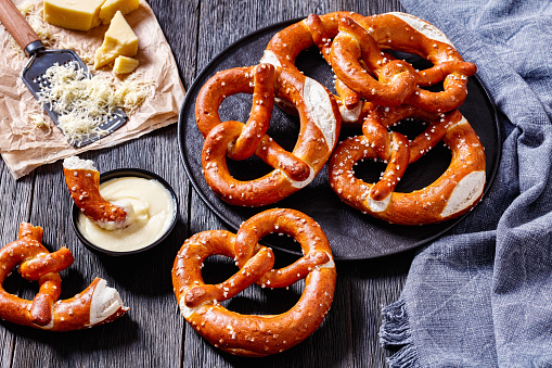 soft pretzels baked in the form of knot and sprinkled with salt on black plate on dark wooden table with cheese sauce, horizontal view from above