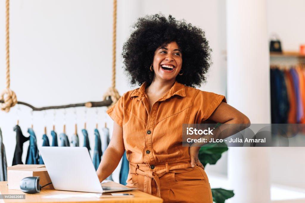 Ethnic small business owner smiling cheerfully in her shop Ethnic small business owner smiling cheerfully while standing in her shop. Happy businesswoman managing her clothing orders on a laptop. Black female entrepreneur running an online clothing store. Small Business Stock Photo