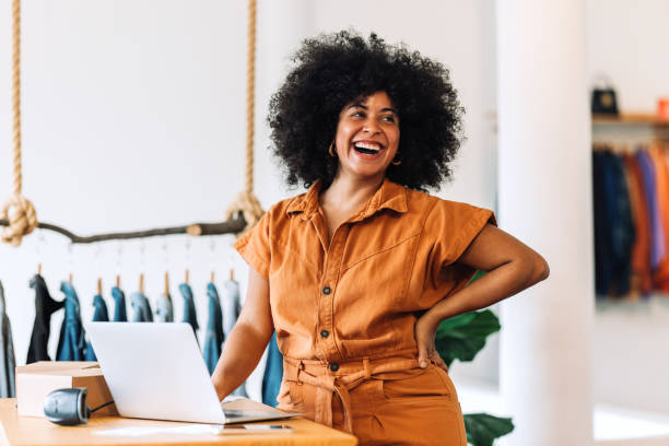 Ethnic small business owner smiling cheerfully in her shop Ethnic small business owner smiling cheerfully while standing in her shop. Happy businesswoman managing her clothing orders on a laptop. Black female entrepreneur running an online clothing store. retail occupation stock pictures, royalty-free photos & images