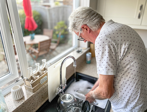 Portrait of a senior man in his late 60s doing the dishes alone in his kitchen at home. Selective focus with room for copy space.