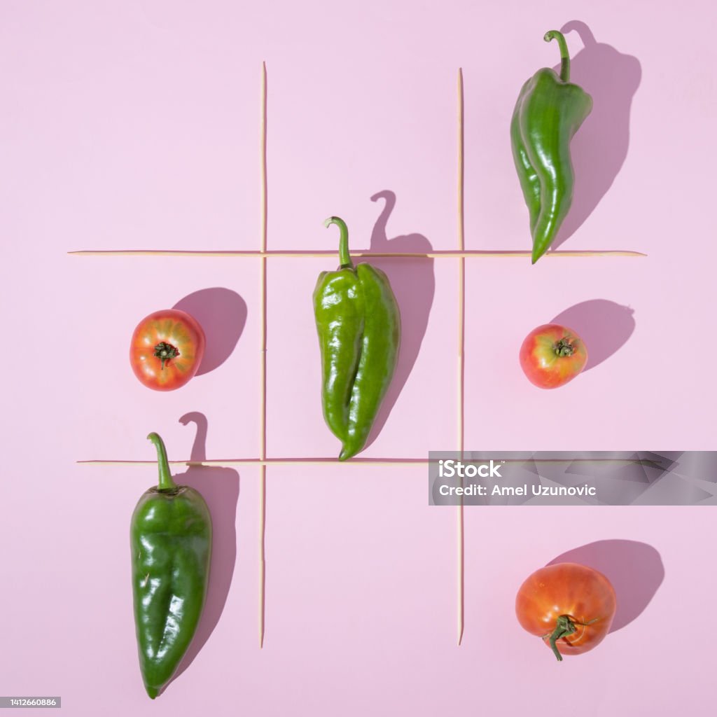Healthy food creative minimal concept. Vegetable x ox game with three organic peppers and three tomatoes on a pastel pink color background and wooden sticks frame. Flat lay design. Abstract Stock Photo