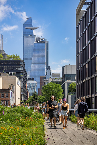 New York, NY, USA - June 24, 2022: Tourists walking on the High Line Park in front of a high rise building with the Edge viewing platform at Hudson Yards against the blue sky. The picture is taken close to West 19th Street