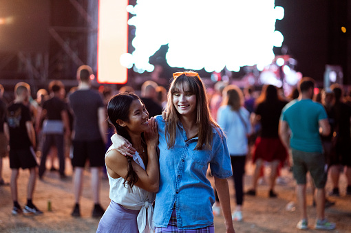 Two girlfriends of Asian and Caucasian ethnicity are hugging, walking, and absorbing the atmosphere at a music festival as the night is falling.
