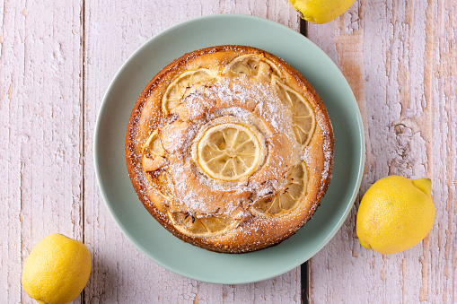 Fresh homemade vegan lemon cake served on a mint colored plate on a white rustic wooden table. Sweet dessert with citrus fruit as a refreshing treat in summer. Top view, close up.