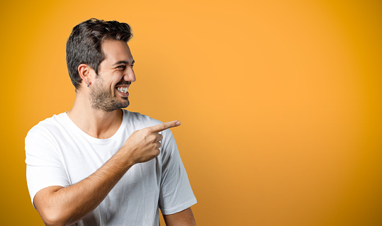 Smiling young man in white t-shirt looks to the side and point finger. Cheerful man isolated on orange background, studio shot for product advertisement.