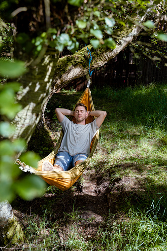 A front view shot of a mid adult man lying down in a hammock with his hands behind his head, he is napping with his eyes closed, feeling relaxed outdoors in Northeastern England.