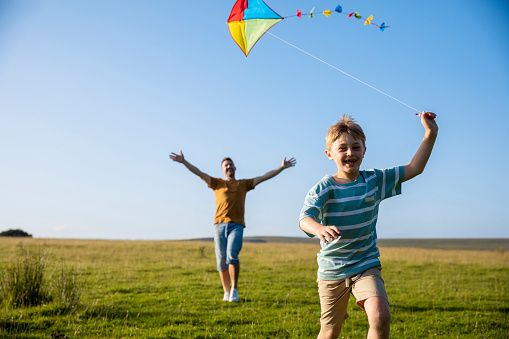A dad and his young son having a great time and running on the grass in a field in the North East of England. The young boy is pulling a multicoloured kite and smiling with glee as they enjoy quality time together.