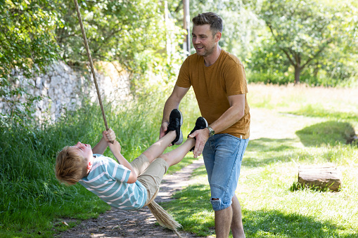 A side-view shot of a young boy and his father smiling and playing on a summers day in the North East of England. The young boy is holding onto a rope and his dad is swinging him.