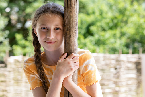 A front-view portrait shot of a young girl smiling and looking at the camera on a summers day in the North East of England. She is holding onto a wooden post.