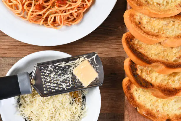 Overhead view of garlic bread on a cutting board and a plate of spaghetti with a cheese grater with grated parmesan cheese.