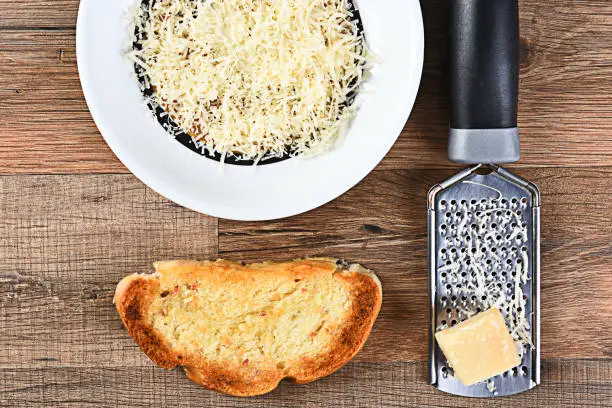 High angle view of a piece of garlic bread and a bowl of grated parmesan cheese with a grater and lump of cheese.