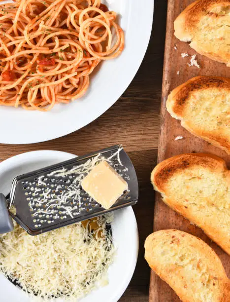 Overhead view of a cheese grater with parmesan cheese on a rustic wood kitchen table. A plate of spaghetti and garlic bread on board are also shown.