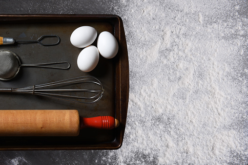 Baking Concept. Top view of a ball of raw bread dough on a baking sheet with a wood rolling pin, eggs, and whisk on a counter top covered with flour.