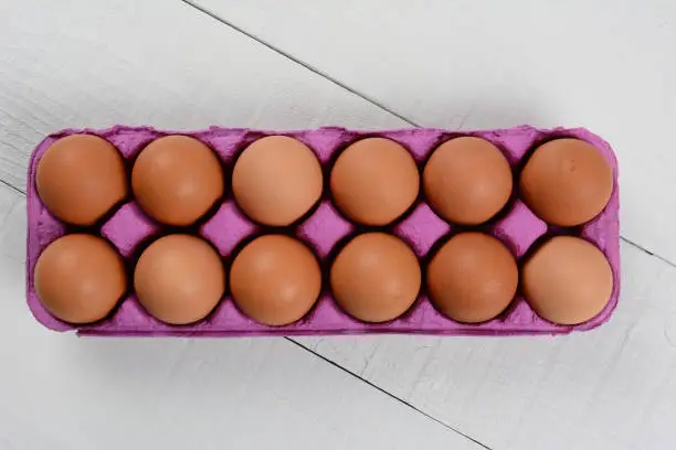 High angle shot of a dozen organic brown eggs in a pink cardboard carton on a rustic white wood table. Horizontal format with copy space.