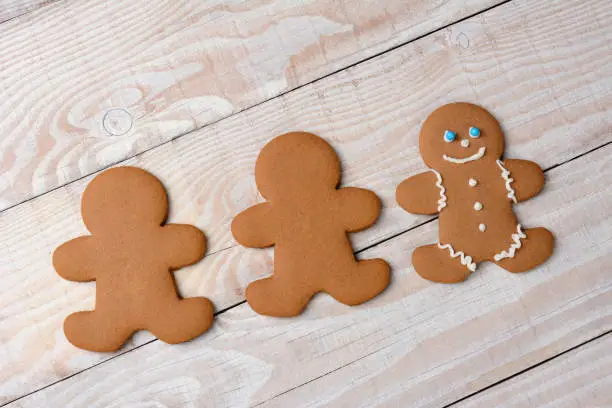HIgh angle view of three gingerbread men on a rustic white kitchen table. Two cookies are  plain and without icing while one is decorated with icing and candies..