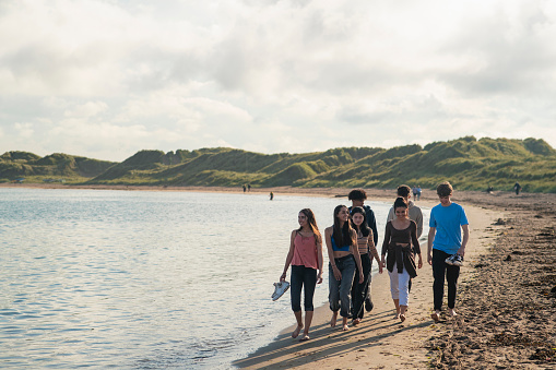 Wide shot of a group of teenage friends relaxing at the beach in Beadnell, North East England. They are talking and walking together along the waters edge.