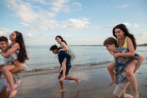 Side view of a group of teenage friends having fun at the beach in Beadnell, North East England. They are running, giving each other piggy backs along the waters edge.