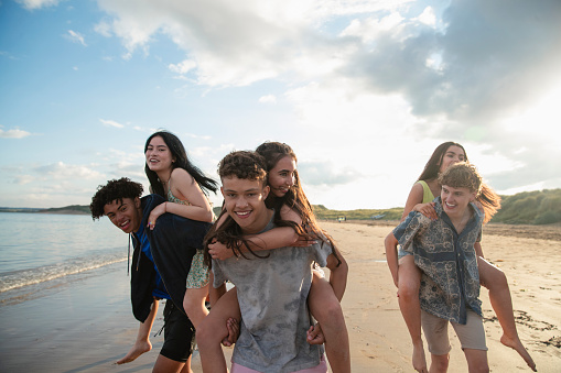 Front view medium shot of a group of teenage friends having fun at the beach in Beadnell, North East England. They are running, giving each other piggy backs along the waters edge. Some of the teens are looking at the camera.