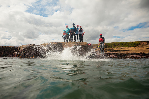 Low angle view of a group of teenagers coasteering, jumping into the sea from a rock in the North East of England at Beadnell. They are all wearing helmets and life jackets, exploring the rocks. The person who has just jumped into the water has made a splash.