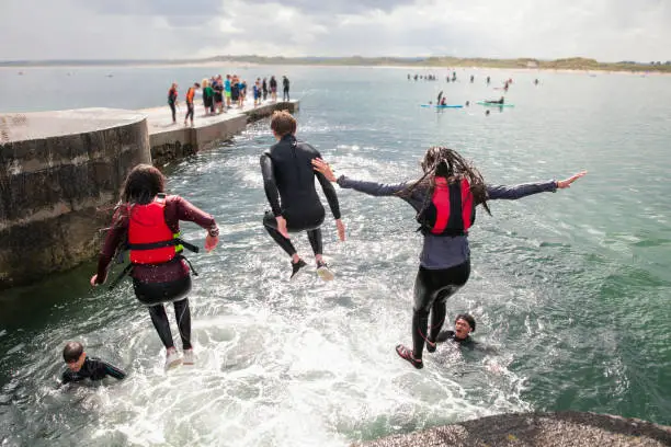 Rear view of a multiracial group of teenage friends wearing wetsuits and life jackets on a jetty on a summers day. There are people paddle boarding in the background.