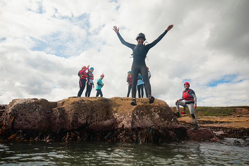Low angle view of a group of teenagers coasteering, jumping into the sea from a rock in the North East of England at Beadnell. They are all wearing helmets and life jackets, exploring the rocks. The person who is jumping has his arms up, making a face.