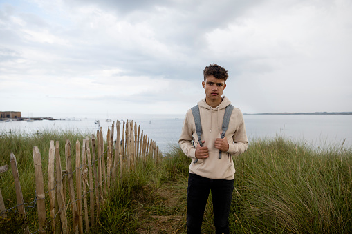 Wide shot of a teenage boy looking at the camera with a serious expression. He is outdoors and is wearing a hoodie and backpack. The ocean is behind him as he explores the coastline in Beadnell, North East of England.