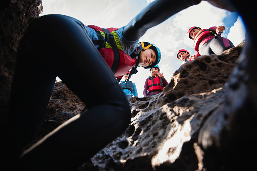 Low angle view of a group of mixed ethnic teens coasteering in the North East of England at Beadnell. They are wearing helmets and life jackets, and one woman is climbing down into a cave while her friends watch from above.