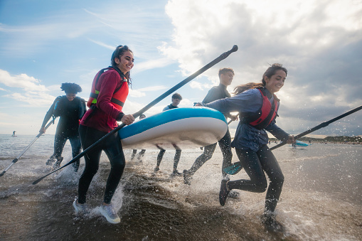 Low angle shot of a group of mixed ethnic and age teens running in the ocean while carrying a multiple person paddle board together in the North East of England at Beadnell. They are wearing wet suits and life jackets to be safe and are laughing, having fun.