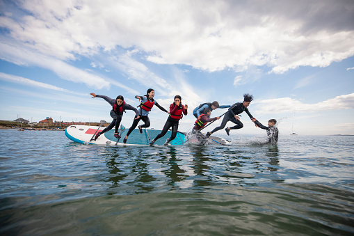 Group of mixed ethnic and age teens jumping off a multiple person paddle board together in the North East of England at Beadnell. They are wearing wet suits and life jackets to be safe and are having fun in the ocean. The teens are holding onto each other as they jump.