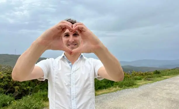 a dark-haired man French Italian or Spaniard shows a heart with his hands he smiles and stands against the backdrop of mountains He is handsome elegant sexy and he has a white shirt High quality photo