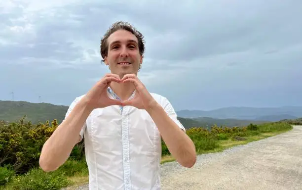 a dark-haired man French Italian or Spaniard shows a heart with his hands he smiles and stands against the backdrop of mountains He is handsome elegant sexy and he has a white shirt High quality photo