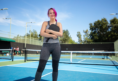 Portrait of white non binary person in their 20s with pink hair holding a pickleball paddle to their chest.