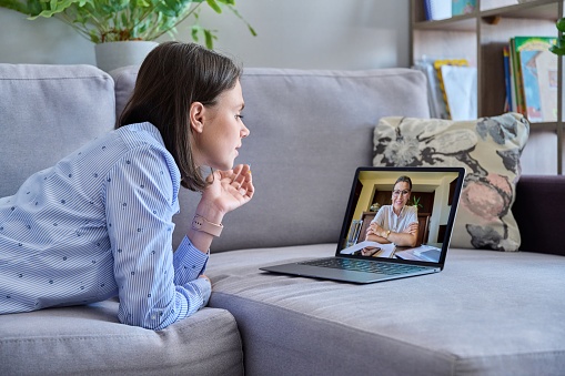 Online therapy meeting of young woman with psychologist. Female lying on couch at home talking with counselor, using laptop for conference call video chat. Technology, psychology, youth, mental health