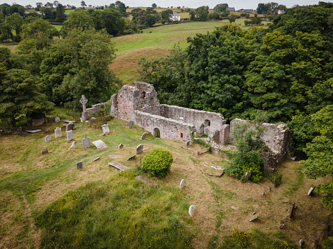 Aerial drone view of the old abandoned church ruins and overgrown graveyard at Layd, Cushendall, Northern Ireland, on a sunny summer day with green grass fields all around, and grass covered headstones, and trees thick with foliage