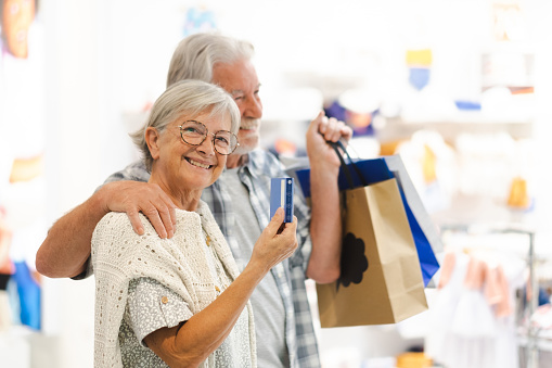 Smiling caucasian senior couple enjoying shopping, man holding shopping bags, the woman takes credit card in hand, consumerism sales customer concept