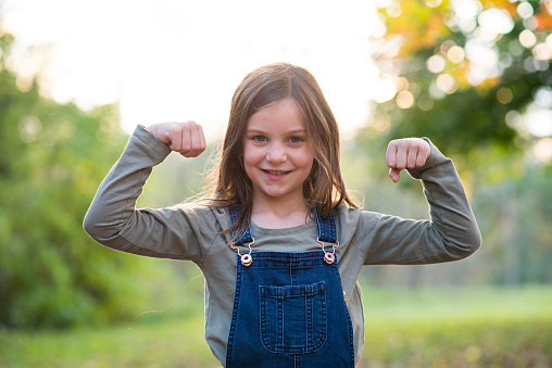 Cute little girl with brown hair and wearing overalls is smiling at the camera as she is flexing her arms and biceps with confidence and strength.