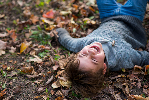 Cute, elementary age boy in a grey knit sweater is laying on the ground on his back in a pile of fallen Autumn leaves and laughing really hard with his eyes closed and a big toothy smile.