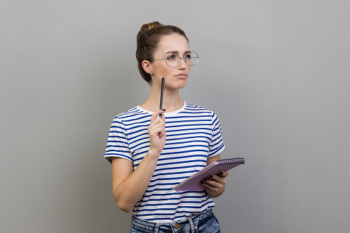 Portrait of woman wearing striped T-shirt making notes in paper notebook, writing business idea, future plans, looking away with thoughtful expression. Indoor studio shot isolated on gray background.