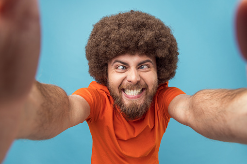 Portrait of crazy silly man with Afro hairstyle in orange T-shirt broadcasting livestream, having fun with subscribers, posing with crossed eyes, POV. Indoor studio shot isolated on blue background.