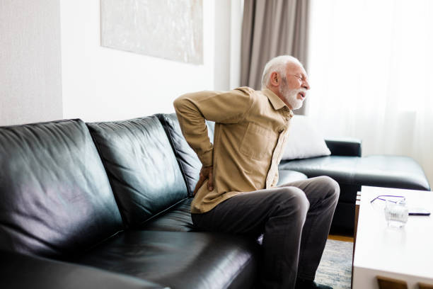 älterer mann fühlt sich unwohl, leidet unter rückenschmerzen, massiert schmerzende muskeln, während er zu hause auf dem sofa im wohnzimmer sitzt. alten- und gesundheitskonzept - arthritis senior adult rheumatoid arthritis sadness stock-fotos und bilder