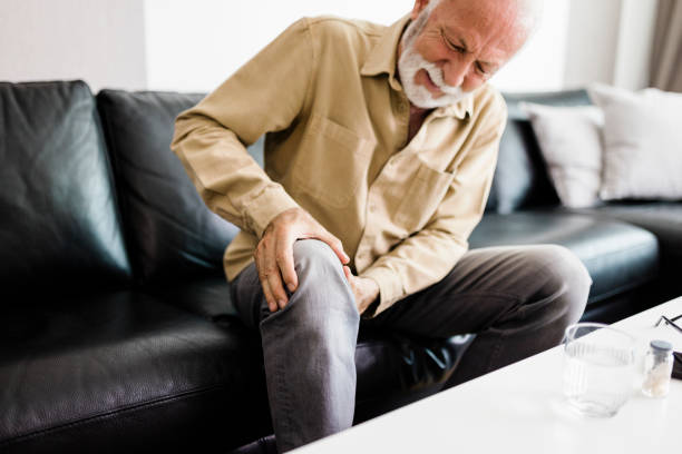 A distraught senior man feeling unwell, suffering from pain in leg while sitting on sofa in the living room at home, Photo of  elderly man sitting on a sofa in the living room at home and touching his knee by the pain during the day. Senior man massaging