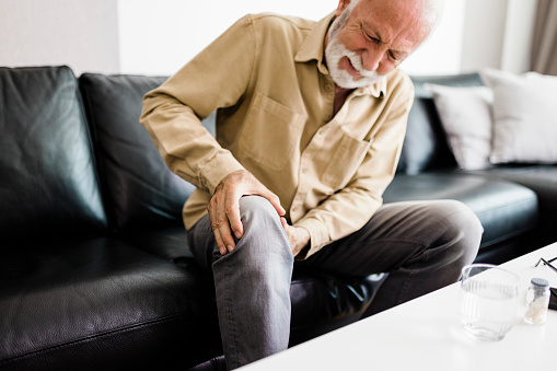 A distraught senior man feeling unwell, suffering from pain in leg while sitting on sofa in the living room at home, Photo of  elderly man sitting on a sofa in the living room at home and touching his knee by the pain during the day. Senior man massaging his painful knee