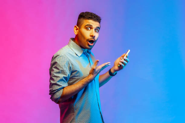 Man in shirt using online service on mobile phone and expressing amazement, looking at camera.