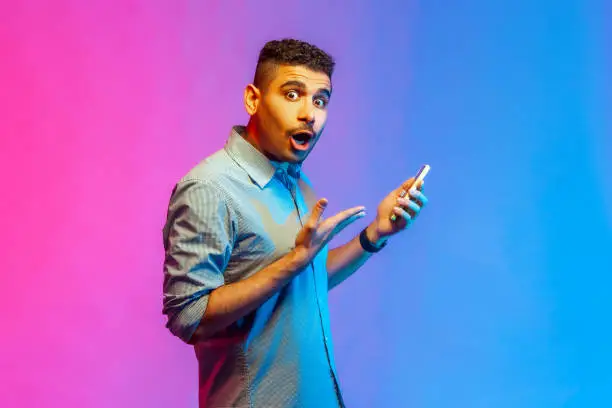 Photo of Man in shirt using online service on mobile phone and expressing amazement, looking at camera.