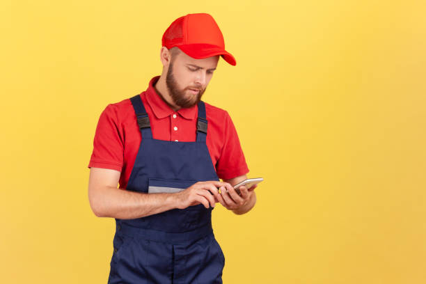 istock Serious handyman in overalls typing on mobile phone using cellphone messenger to accept online order 1412729555