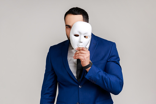 Bearded man holding white mask, covering face, standing with serious expression, multiple personality disorder, wearing official style suit. Indoor studio shot isolated on gray background.