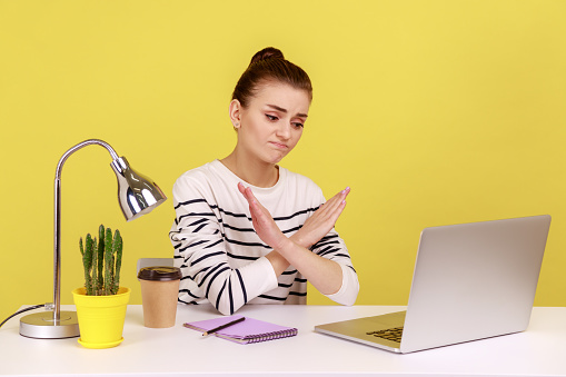 Upset sad woman manager sitting at workplace crossing hands showing x sign, looking at laptop screen with frowning face. Indoor studio studio shot isolated on yellow background.