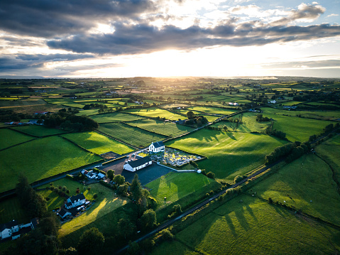A beautiful aerial sunset over the County Antrim countryside, in summer, with agricultural land and sun beams catching the grass in fields, and Killymurris church illuminated in the distance, taken at Glarryford, County Antrim, Northern Ireland