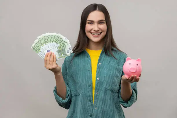 Photo of Smiling affirmative woman holding big sum of euro banknotes and piggy bank, profitable investment.